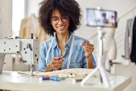 Happy African-American clothes designer shoots tutorial for followers in spacious workshop