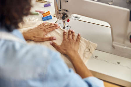 African-American worker sews white fabric with machine tool at table in studio