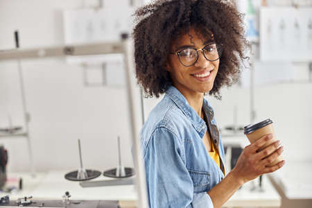 African-American seamstress with glasses and earring in nose holds cup near sewing machine in...