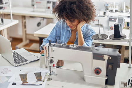 African-American woman clothes designer draws sketches at table with sewing machine and laptopの写真素材