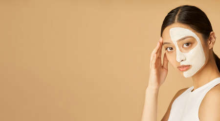 Website header of Studio shot of young woman with facial mask applied on half of her face looking at camera, receiving spa treatments isolated over beige backgroundの写真素材