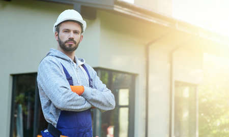 Portrait of young bearded male engineer in hard hat looking at camera, posing outdoors while working...