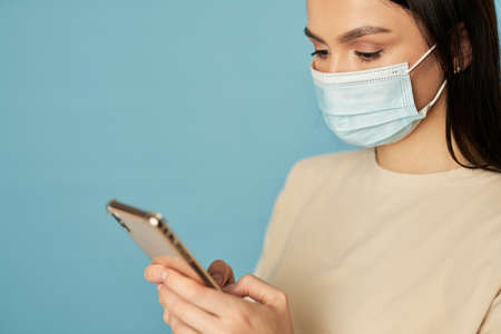 Young lady in mask using smartphone and looking at screen in studio