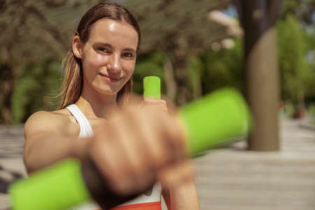 Woman does sports exercises withcolorful dumbbells in city park on nice summer day