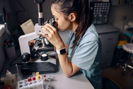 Asian woman lab assistant looks into microscope at workplace in hospital laboratory