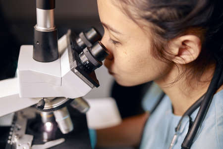 Asian lady doctor looks into contemporary microscope researching sample on slide in hospital