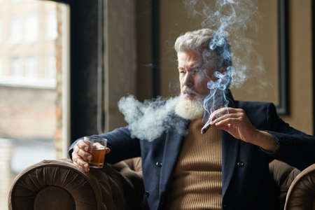 Elegant mature businessman smoking cigar with fume and holding a glass of whisky while relaxing in a chair in loft interiorの写真素材