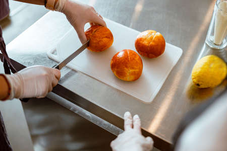 Worker in gloves cuts fresh bun with bread knife with colleague in bakeryの写真素材