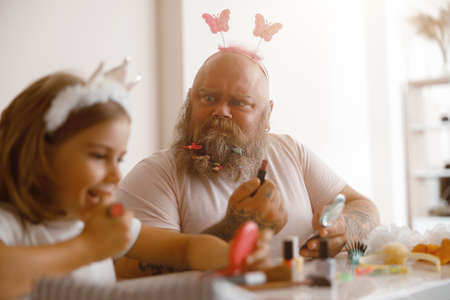 Bearded man looks at little girl applying lipstick at table in living roomの写真素材