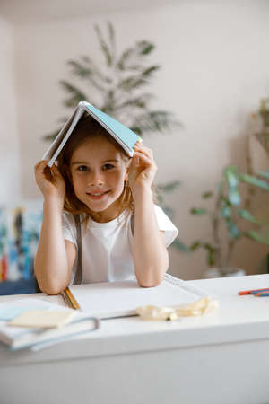 Happy little girl holds notebook on head sitting at table in light room