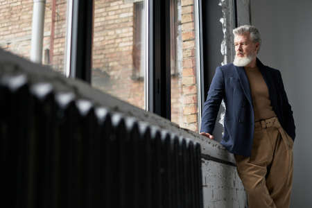 Stylish mature man in business casual wear looking out a large window, being thoughtful while...