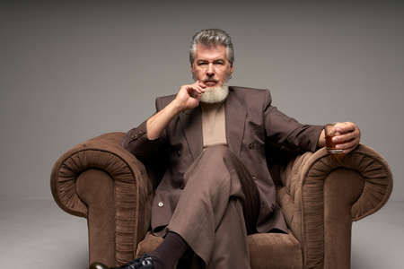 Thoughtful mature businessman with beard wearing elegant suit looking at camera, holding a glass of...