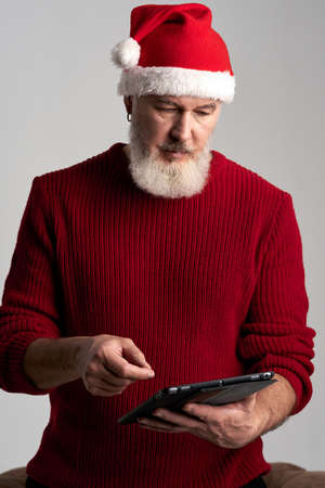 Modern Santa. Focused middle aged man with beard wearing Christmas hat and red sweater using digital tablet while standing isolated over gray backgroundの写真素材