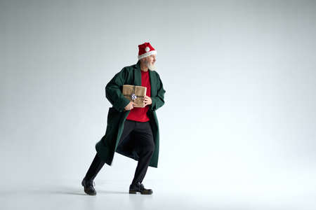 Full length shot of cool middle aged man with Christmas hat wearing casual outfit looking away, holding gift box while posing isolated over light gray backgroundの写真素材