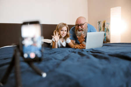 Cheerful man and pretty daughter bloggers with laptop wave hands to greet followers at homeの写真素材