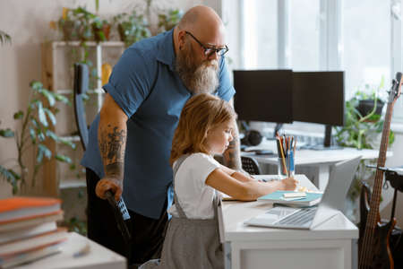Bearded plump father stands looking at little girl doing hometask at homeの写真素材
