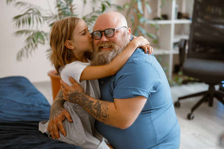 Little girl hugs and kisses smiling bearded plump father sitting on bed at homeの写真素材