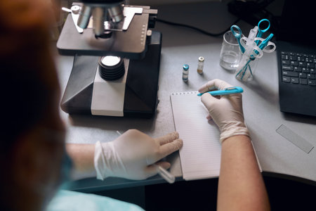 Lab tech in latex gloves writes marks in notebook near microscope at deskの写真素材