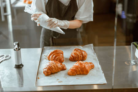 Employee holds confectioner sleeve with delicious cream to decorate croissants in bakery shopの写真素材