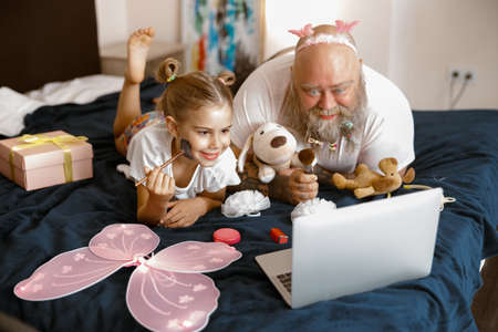 Little girl and father with cosmetic brushes watch beauty blogger video on bed at homeの写真素材