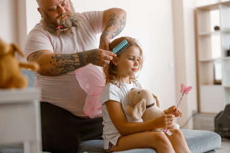 Daddy with scrunchies in beard brushes hair of cute little daughter at homeの写真素材