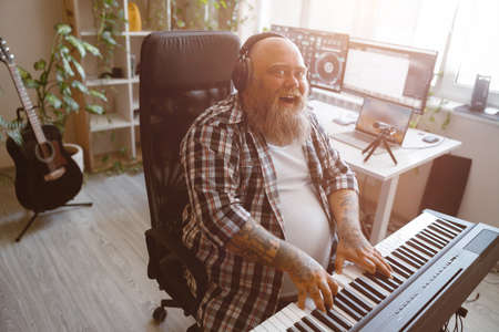 Joyful obese man artist plays music on synthesizer near table with computer monitors in studioの写真素材