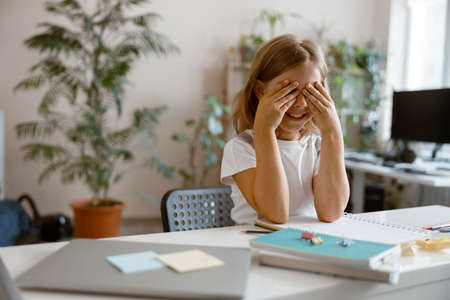 Little child closes eyes by hands sitting at table with supplies and laptop in roomの写真素材