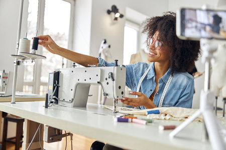 Happy African-American woman puts coil onto sewing machine shooting new video in studio