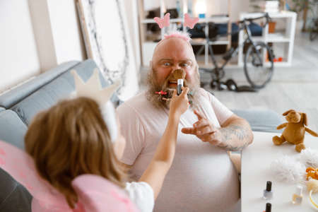 Daughter with princess crown applies powder onto nose of shocked father in living roomの写真素材