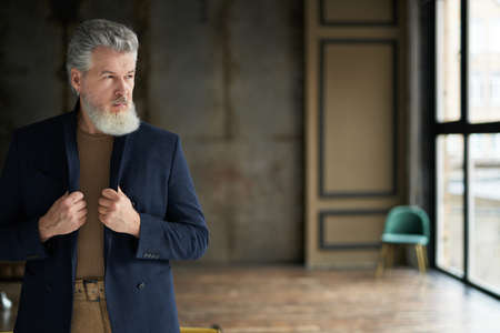 Stylish serious handsome middle aged man looking away, adjusting his suit while posing indoors