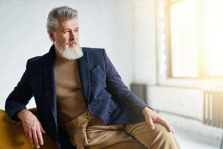 Handsome middle aged business man with a beard looking away while sitting on sofa indoorsの写真素材