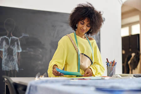 African-American woman with tape measure and belt bag looks at paper at sewing class in studio