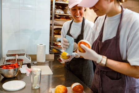 Happy woman with grater and lemon and colleague filling bun with cream in bakery