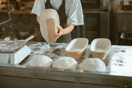 Woman holds large dish for bread at table with raw dough loafs in bakery