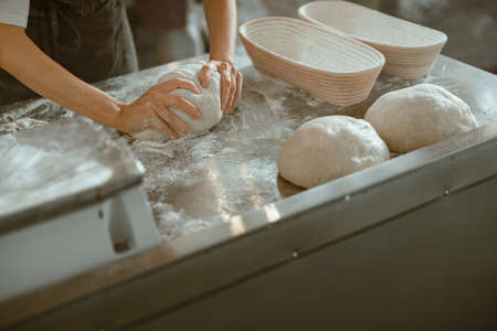 Worker kneads dough making bread at metal table in modern craft bakery shop