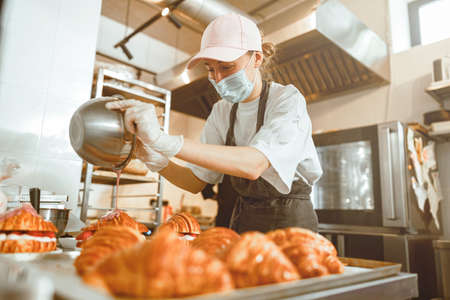 Lady in mask pours pink icing onto croissant at countertop in bakeryの写真素材