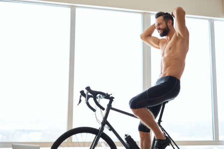 Cheerful young man with athletic body using bicycle trainer at homeの写真素材