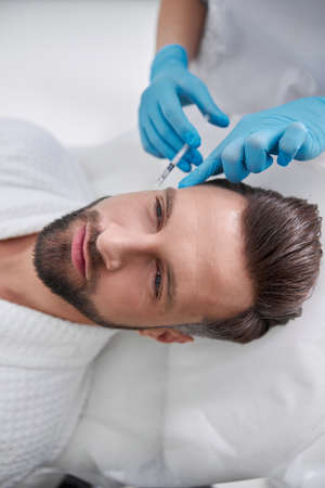 Cosmetologist holds finger on mature man temple after injection of filler for skin lifting in beauty salonの写真素材