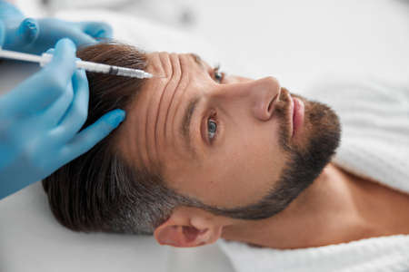 Doctor cosmetologist does injection of lifting filler in wrinkled forehead of mature man in clinicの写真素材