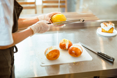 Lady grates lemon zest onto tasty buns with cream at metal counterの写真素材