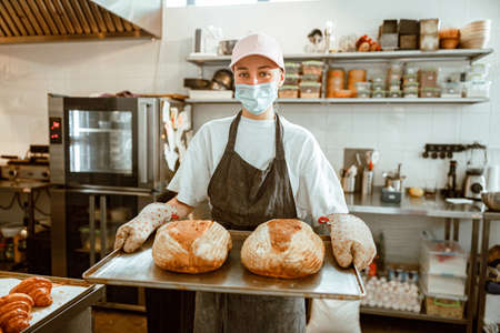 Woman with medical mask holds tray with freshly baked breads in craft workshopの写真素材