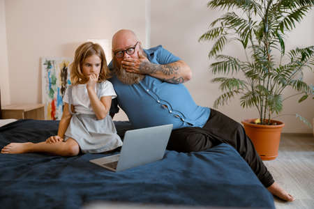 Scared man and little girl watch video via modern computer sitting on bed at homeの写真素材