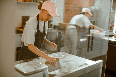 Pretty woman cleans countertop from flour after preparing dough in bakery workshopの写真素材