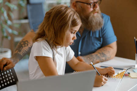 Pretty little girl writes in copybook sitting nera laptop doing homework with daddy at home