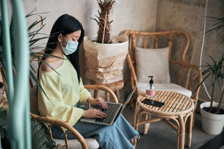 Asian woman in protective mask sits in cafe at laptop