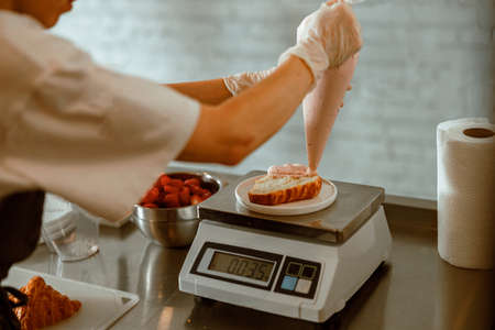 Confectioner applies pink cream onto croissant on scales in craft bakery shopの写真素材
