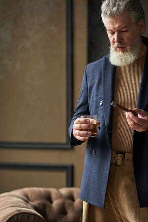 Cropped shot of serious mature man with beard looking at cigar while holding it and glass of whisky,...