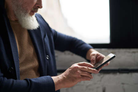Cropped shot of bearded middle aged man in business casual wear using his smartphone while sitting by a window in modern loft interiorの写真素材