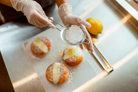 Lady pours powdered sugar onto round buns with cream at table in bakeryの写真素材