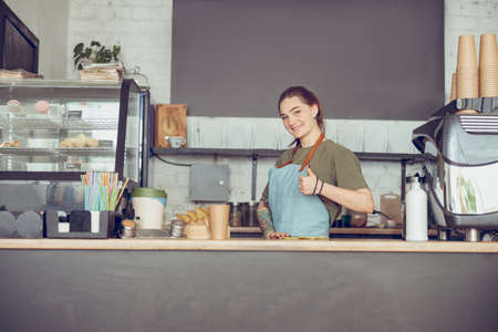 Joyful barista standing behind counter and giving thumbs upの写真素材